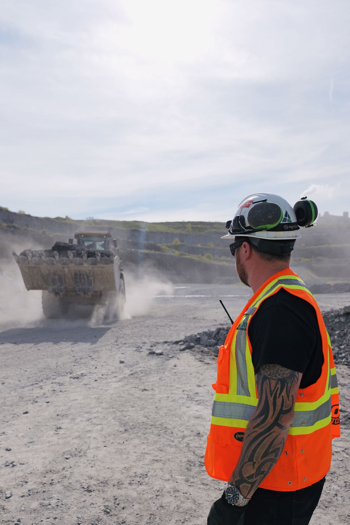 Crew member with a radio in a quarry as a loader passes through dust
